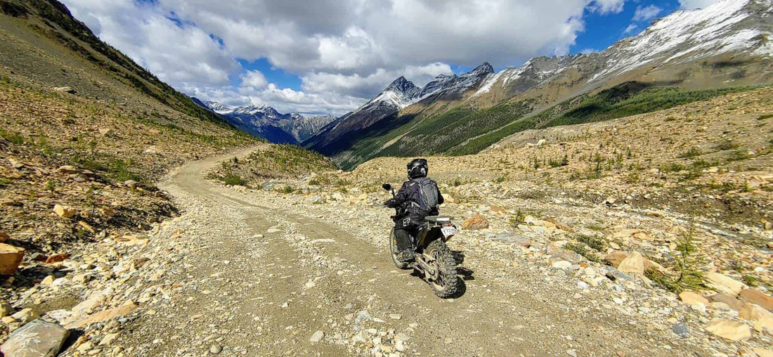 Rider on a loaded adventure motorcycle on a rocky alpine gravel pass near Invermere with snow-dusted peaks ahead