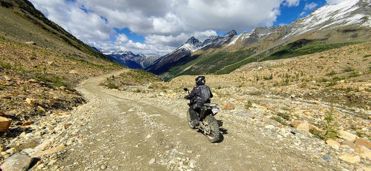 Rider on a loaded adventure motorcycle on a rocky alpine gravel pass near Invermere with snow-dusted peaks ahead