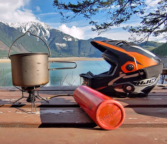 MSR Whisperlite International stove and titanium pot beside an orange moto helmet on a lakeside table