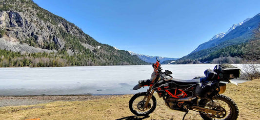 KTM adventure motorcycle parked beside a frozen Trout Lake with snowy Kootenay mountains behind