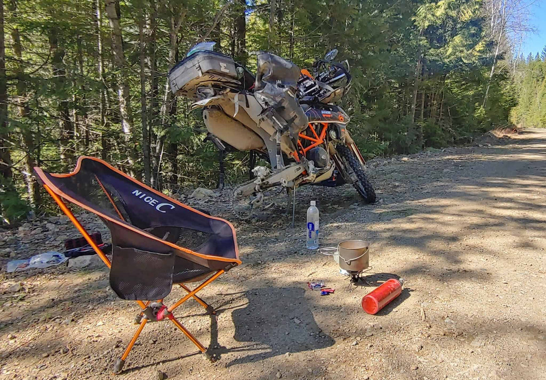 KTM adventure bike on its side stand at a forest road camp with a chair, stove and tire repair kit