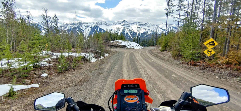 Rider point of view over an adventure bike on a snow lined gravel road below snowy Kootenay peaks