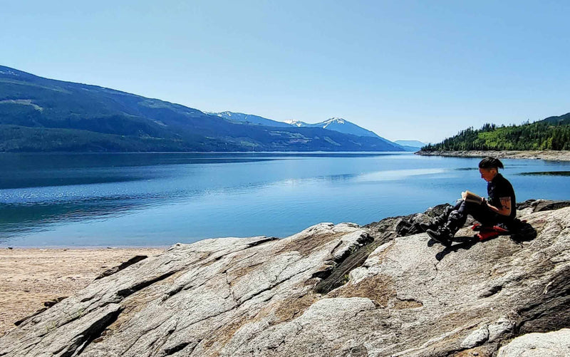 Rider sitting on a granite shoreline reading beside a calm West Kootenay lake ringed by mountains