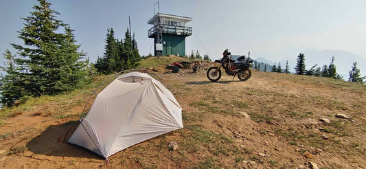 Solo moto camping kit with an ultralight tent and adventure bike beside a backcountry fire lookout tower
