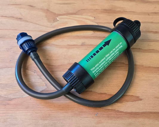 Green ultralight outdoor water filter with hose attached, coiled on a wood surface for moto camping