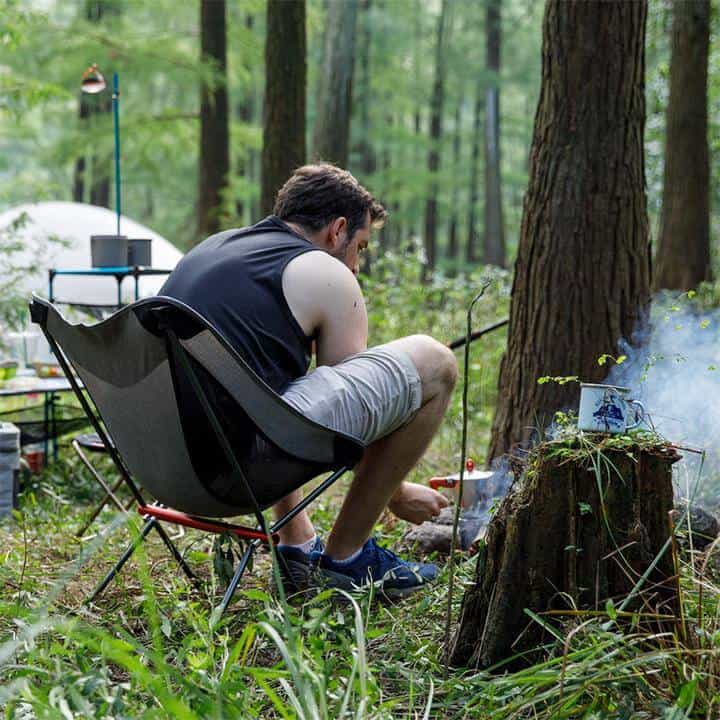 Naturehike collapsible camp chair set up in a forest campsite with a man tending a fire nearby