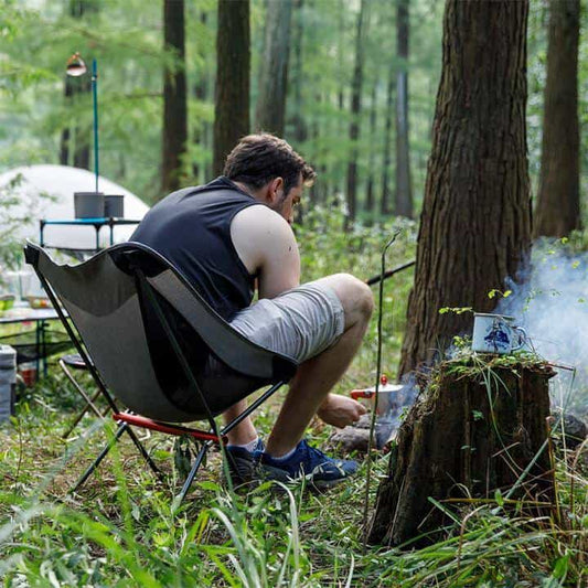 Naturehike collapsible camp chair set up in a forest campsite with a man tending a fire nearby