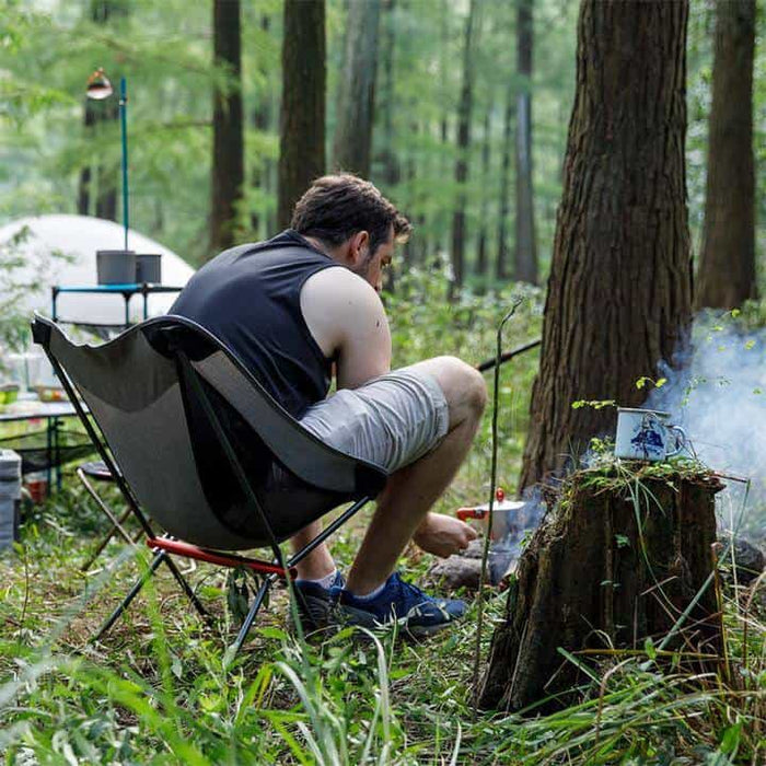 Naturehike collapsible camp chair set up in a forest campsite with a man tending a fire nearby