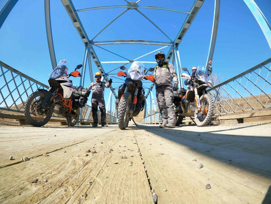 Three adventure riders and KTM bikes posing on a wooden deck bridge under a blue steel truss