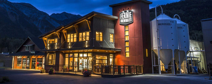 Fernie Brewing Co. building lit up at dusk with Kootenay mountains behind and Meridian grain silos alongside
